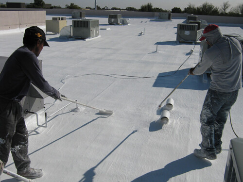 Two Sunland roof coating speicalists coating a flat roof with elastomeric paint from Sunland Coatings MFG. One man is spraying the paint and one is spreading the paint on a flat white roof.